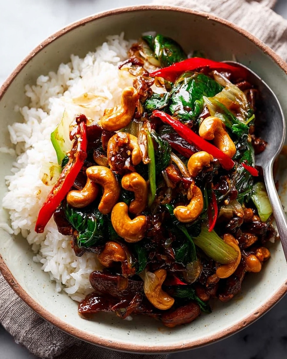 A white bowl filled with two layers: the bottom layer is fluffy, white cooked rice with defined grains, and the top layer is a mix of glossy, golden-brown cashew nuts, sliced bright red chili peppers, caramelized brown onions, and wilted dark green leafy vegetables. The ingredients on top have a shiny, slightly oily texture, showing they are cooked in a sauce. A copper spoon is placed on the right side inside the bowl, resting on the rice. The bowl is on a folded brown cloth, set against a white marbled surface. photo taken with an iphone --ar 4:5 --v 7