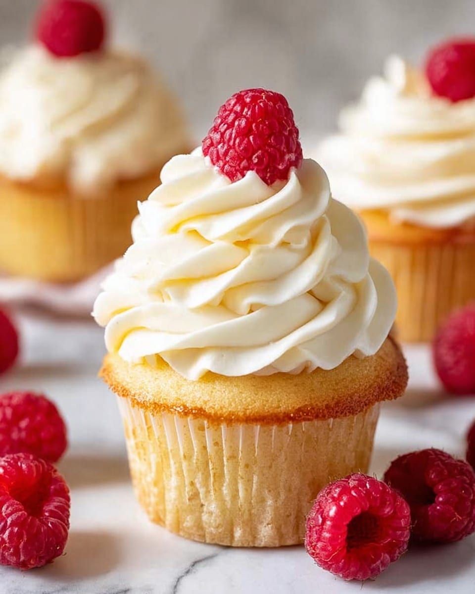 A single golden yellow cupcake sits in the center with its white paper liner peeled back, showing its soft texture. On top of the cupcake is a tall swirl of light, creamy white frosting with smooth, ruffled folds, crowned by a bright red raspberry. Around the cupcake, several fresh raspberries rest on the white marbled surface, adding pops of deep red. In the blurred background, other similar cupcakes are visible with their creamy frosting peaks. The scene is bright and clean, focused closely on the main cupcake. photo taken with an iphone --ar 4:5 --v 7