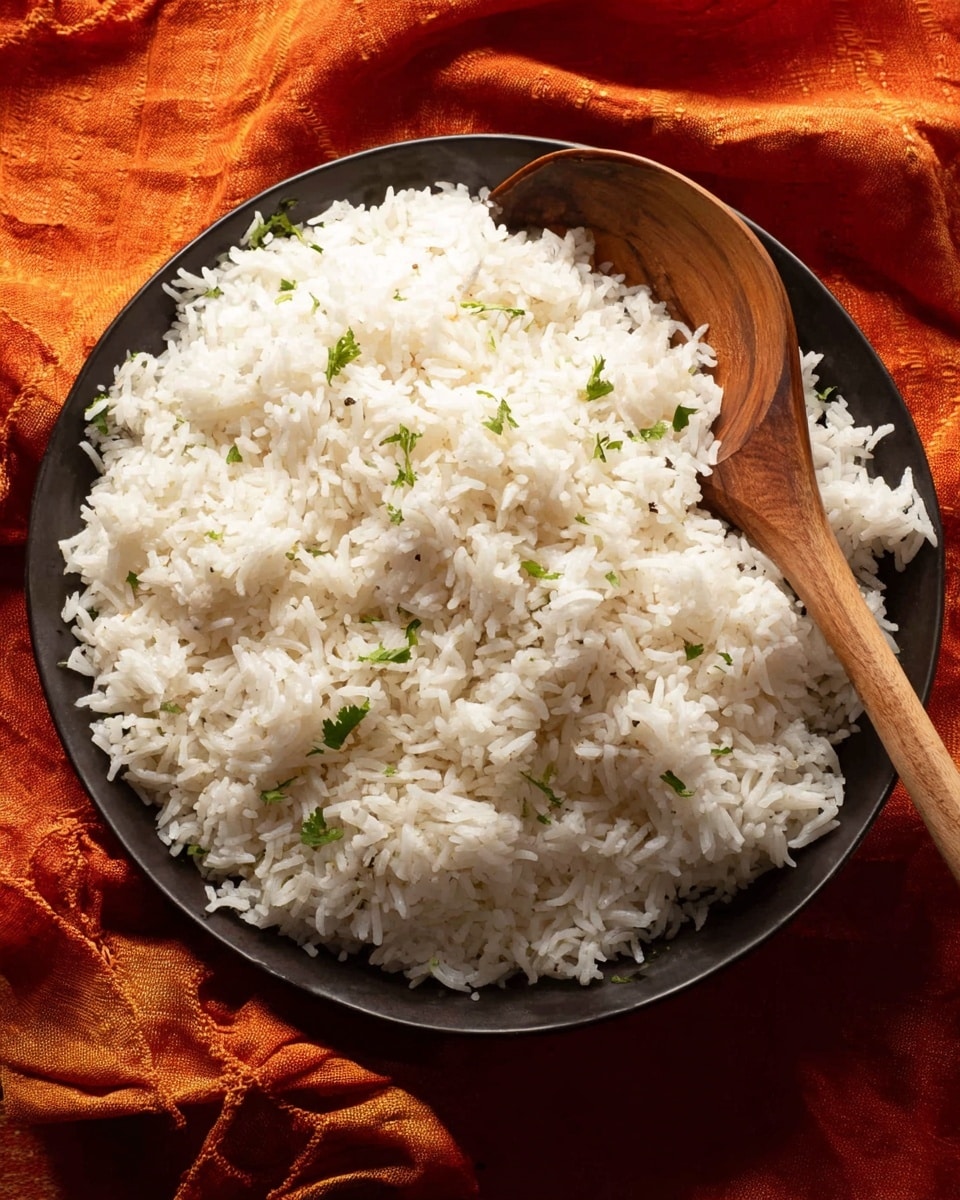 A single layer of fluffy, white cooked rice sits spread evenly across a deep round dark bowl, with scattered small pieces of fresh green herbs adding contrast on the surface. A smooth brown wooden spoon rests on the right side inside the bowl, its handle extending outward. The bowl is placed on a crinkled, vibrant red-orange fabric that adds warmth and texture to the scene. Photo taken with an iphone --ar 4:5 --v 7