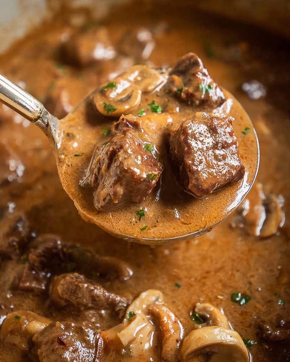A close-up view of a spoon lifting thick creamy brown gravy filled with small, tender-looking meat chunks and sliced mushrooms. The sauce is smooth and glossy, coating the ingredients fully with a few green herb bits scattered on top for color. The background shows more of the same rich sauce and meat mixture, all set against a white marbled texture. photo taken with an iphone --ar 4:5 --v 7