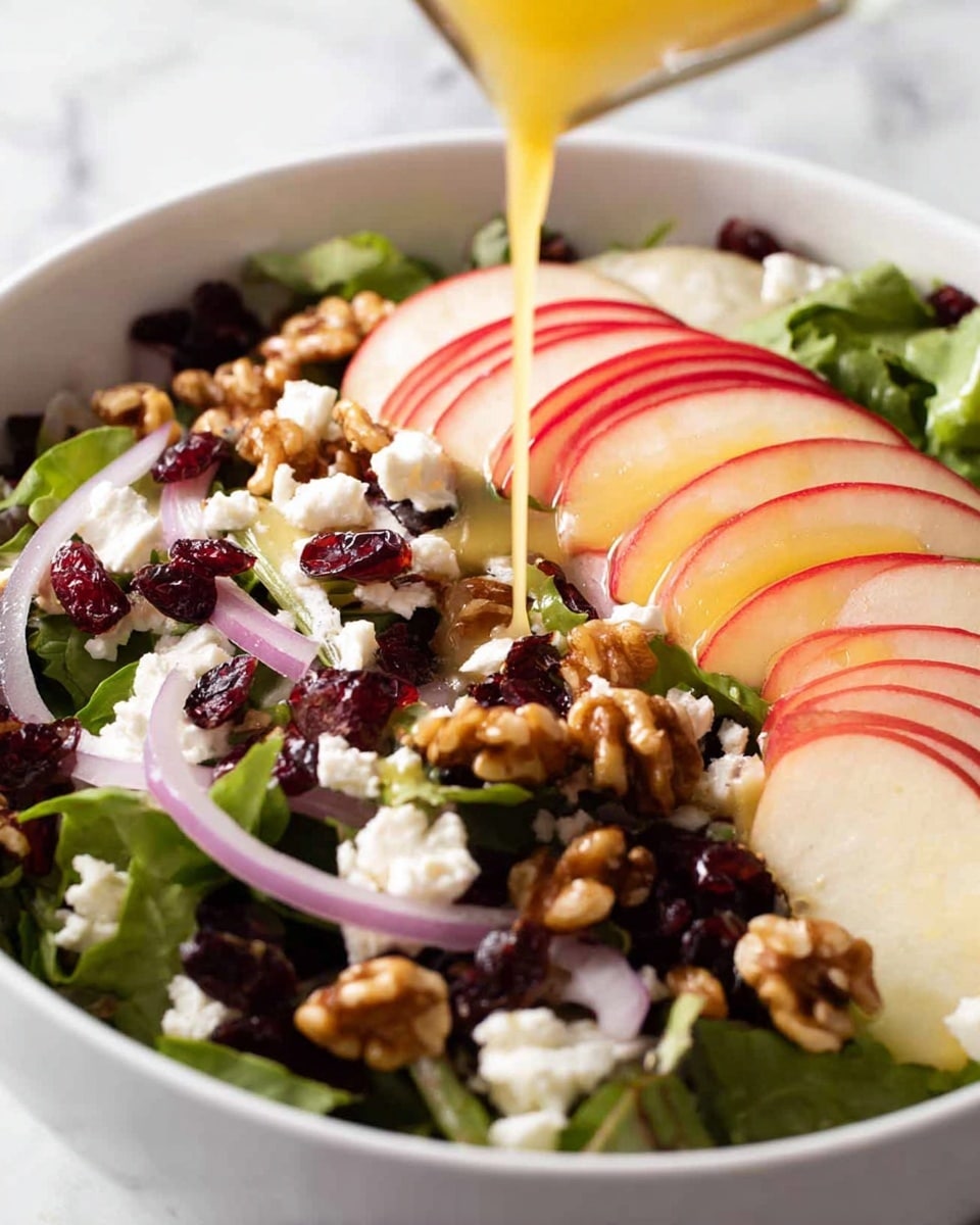 A white bowl filled with a salad shows a base layer of green leafy vegetables mixed with thin purple onion slices. On top, there are neat rows of thinly sliced red-skinned apples with white flesh, sprinkled with small white crumbles of cheese. Scattered among the apple slices and greens are dark red dried cranberries and chunks of golden brown walnuts. A woman's hand pours a smooth, light yellow dressing over the center of the salad, adding a shiny glaze to the apple slices and nuts. The bowl sits on a white marbled textured surface. photo taken with an iphone --ar 4:5 --v 7