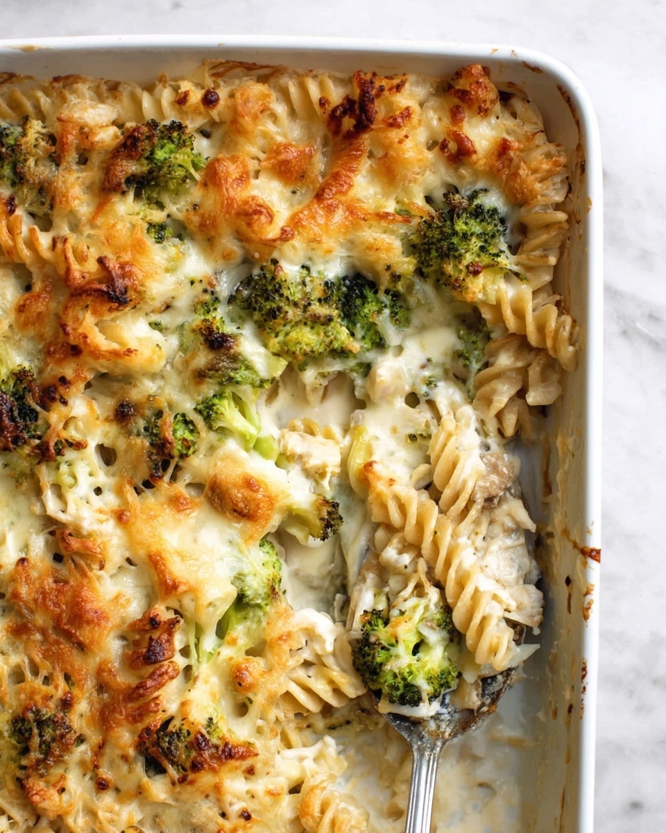 A white baking dish filled with a baked pasta casserole showing three main layers: at the bottom, there are some pieces of cooked chicken with herbs mixed in, followed by spiral pasta in a light yellow color, and topped with bright green broccoli florets covered in a creamy white sauce with melted and browned cheese, giving a bubbly and slightly crispy texture. A metal serving spoon is scooping a portion from the dish, revealing a creamy inside with visible pasta and broccoli in the white marbled background. photo taken with an iphone --ar 4:5 --v 7