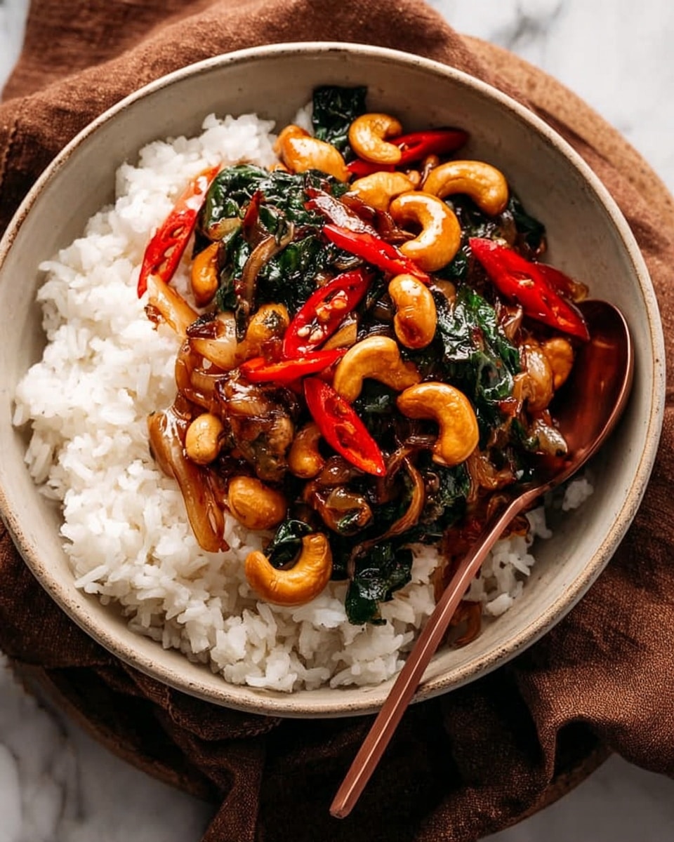 A close-up image of a white bowl filled with two main layers: the bottom layer is plain white rice with a soft and fluffy texture, and the top layer is a mix of glossy, dark brown stir-fried cashew nuts, shiny green leafy vegetables, thinly sliced translucent onions, and bright red chili slices scattered throughout. The bowl sits on a towel over a white marbled surface, and the food looks rich and savory with a slight oil sheen. A metal spoon is partially visible inside the bowl. Photo taken with an iphone --ar 4:5 --v 7
