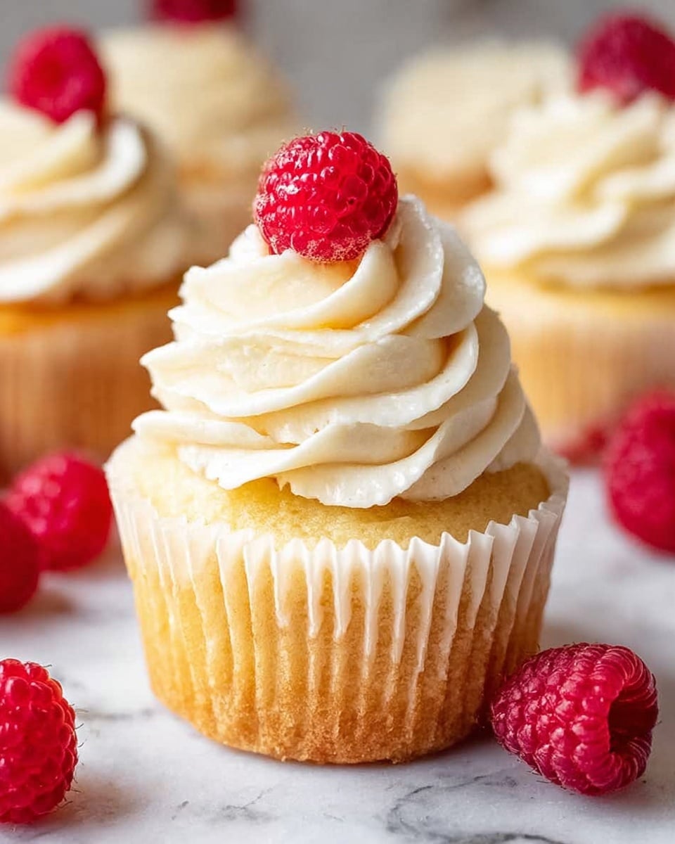 The image shows a single cupcake with its paper wrapper peeled down around the base, revealing a light golden yellow cake layer. On top of the cupcake is a tall swirl of creamy white frosting, soft and textured with gentle waves and peaks. A fresh red raspberry is placed at the very top of the frosting as a bright, colorful accent. Around the cupcake on a white marbled surface are several more raspberries. In the blurred background, there are two more cupcakes with similar frosting. photo taken with an iphone --ar 4:5 --v 7