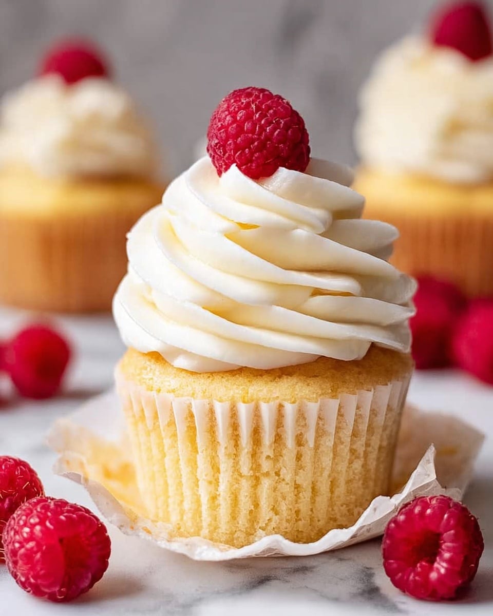 A single golden yellow cupcake sits with its white paper wrapper peeled down to show the soft, moist cake. On top, there is a tall swirl of smooth, white cream frosting, shaped in soft peaks, crowned with a bright red raspberry. Around the cupcake on the white marbled surface, several fresh raspberries are scattered, adding vibrant color to the scene. In the blurred background, more cupcakes with similar frosting are visible, all arranged on the same white marbled texture. photo taken with an iphone --ar 4:5 --v 7
