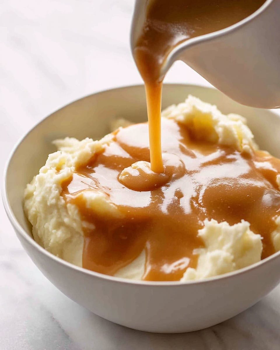 A close-up view of creamy mashed potatoes in a white bowl, with smooth light brown gravy being poured over the top, creating a glossy, thick layer spreading across the fluffy white base. The mashed potatoes have a soft, slightly textured surface with gentle peaks surrounding the gravy pool in the center. The background is a white marbled texture, highlighting the warm, inviting colors of the dish. photo taken with an iphone --ar 4:5 --v 7