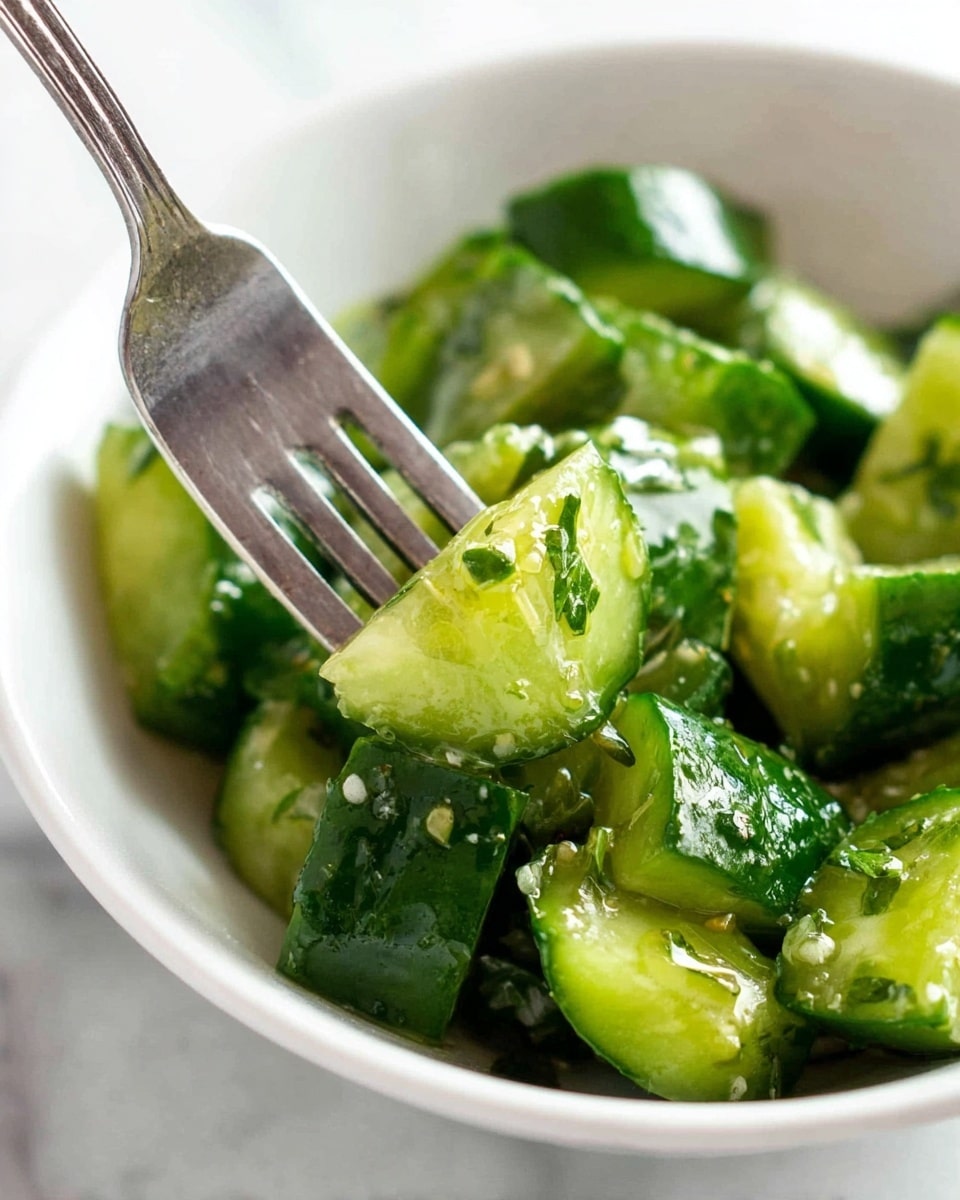 A close-up image of a white bowl filled with bright green cucumber pieces, some crushed and glistening with a light dressing. The cucumber chunks have dark green skin and pale green, watery flesh. Small bits of chopped herbs are mixed throughout. A silver fork presses down on one cucumber piece, showing a bit of soft texture inside. The background is a white marbled surface. photo taken with an iphone --ar 4:5 --v 7