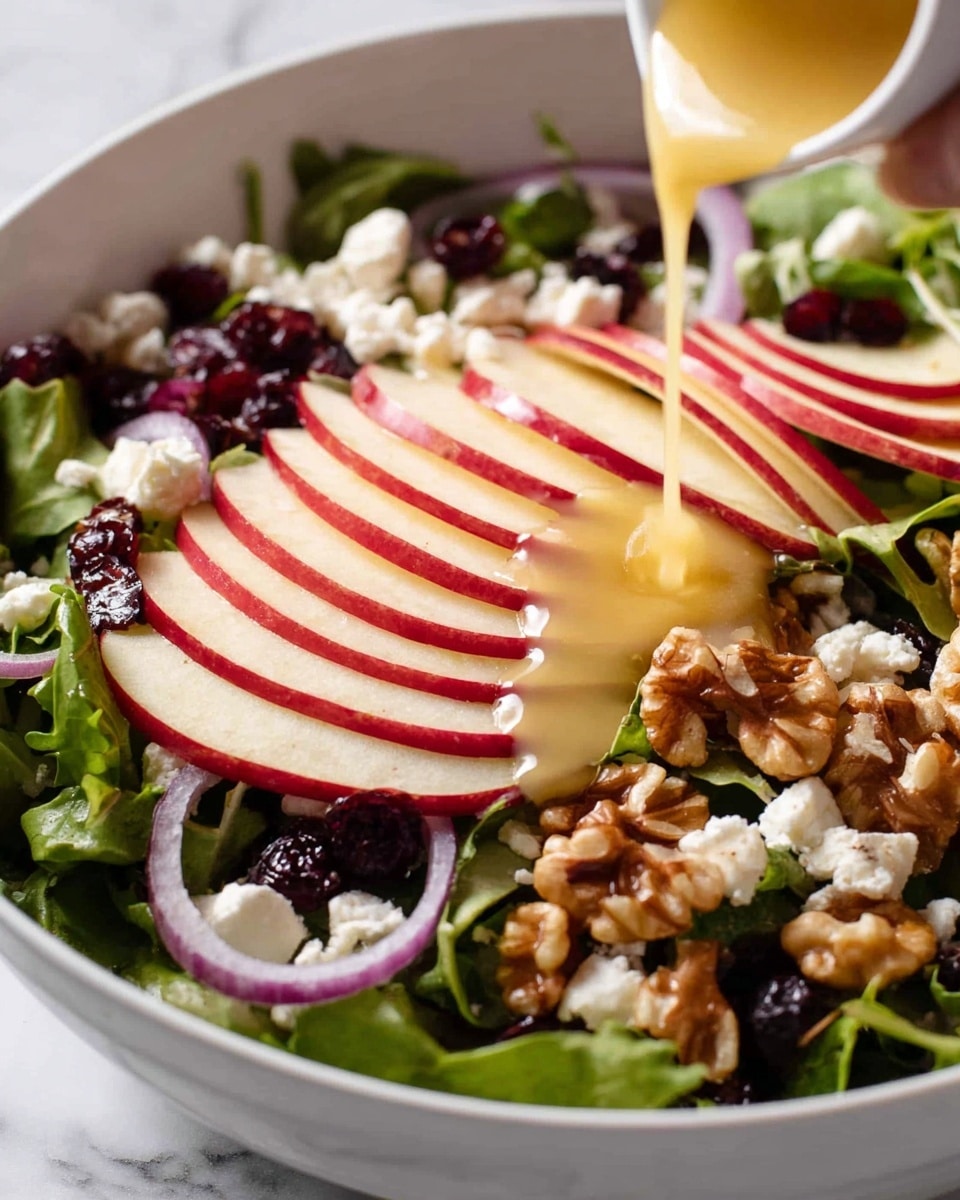 A white bowl filled with a fresh salad on a white marbled surface. The bottom layer of the salad is green leafy greens with a soft texture, topped with thin slices of red apple arranged in curved rows on one side. Scattered over the salad are small, dark red dried cranberries and crunchy walnut pieces with a light brown color. Thin slices of light purple onion are layered among the greens, adding a slight contrast. Small white crumbles of cheese are sprinkled evenly on top. A yellow dressing is being poured over the salad from a glass container, landing mostly on the apple slices and nuts. photo taken with an iphone --ar 4:5 --v 7