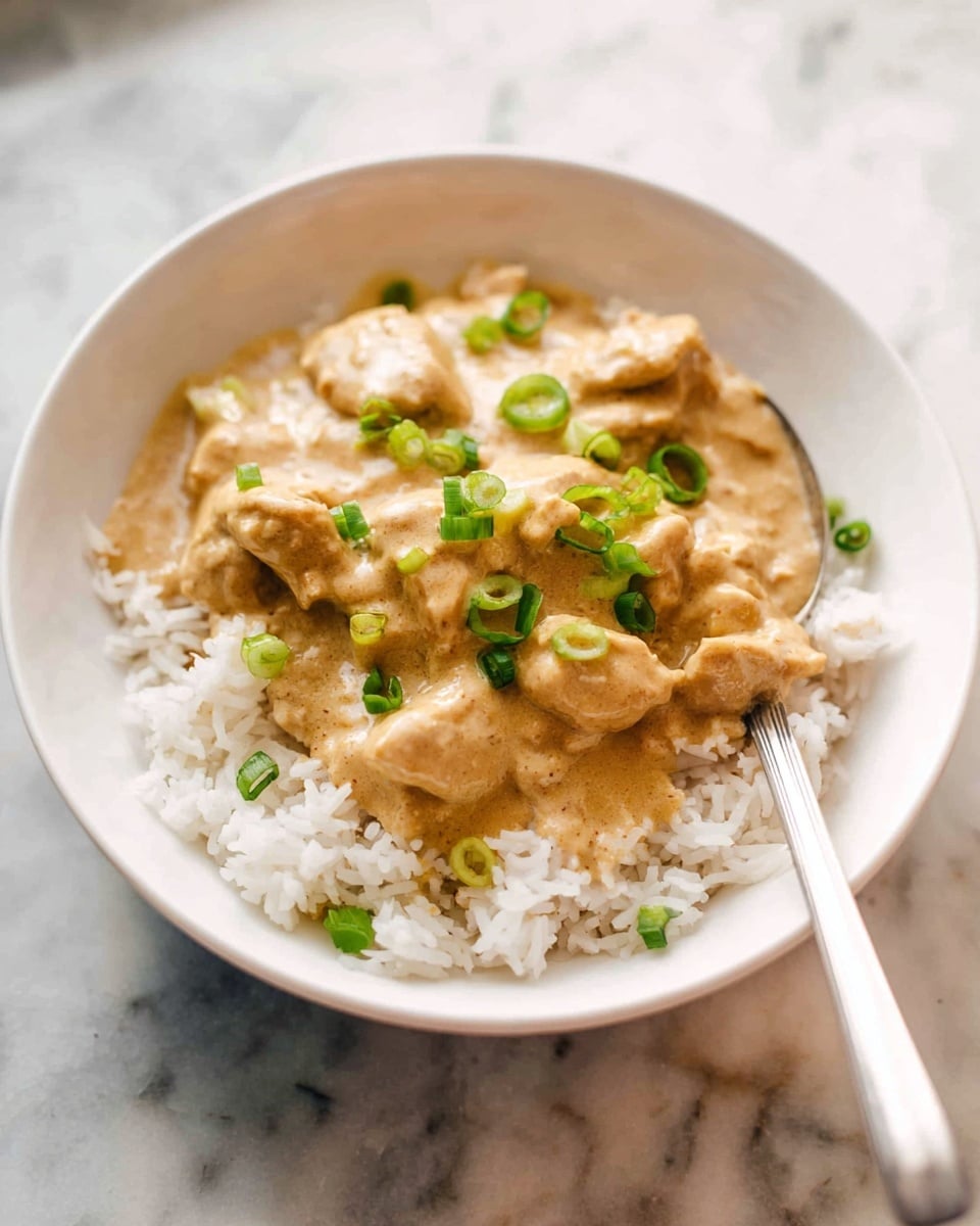 A white bowl filled with a base layer of fluffy white rice, topped with pieces of chicken covered in a thick, light brown creamy sauce, garnished with chopped green onions scattered on top. A silver spoon rests inside the bowl, slightly touching the sauce. The bowl is placed on a white marbled surface with soft, natural lighting highlighting the textures and colors. photo taken with an iphone --ar 4:5 --v 7