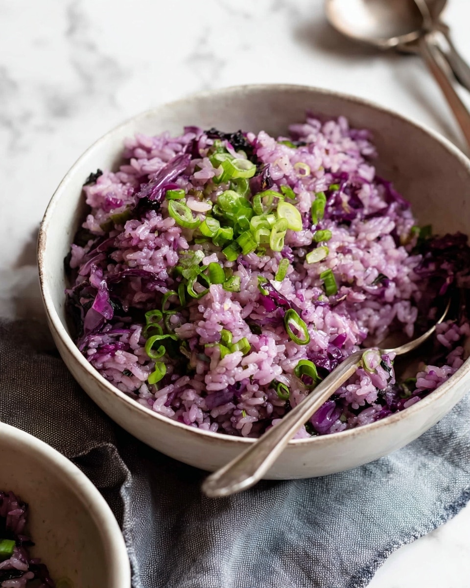 A bowl filled with two layers: the base layer is soft rice mixed with purple cabbage, giving a mix of light pink and deep purple colors with a slightly moist texture. On top, small green onion rings are scattered, adding a fresh green contrast. The bowl is white, round, and simple, with a silver spoon resting inside it towards the right. The bowl sits on a light beige cloth over a white marbled surface. Photo taken with an iphone --ar 4:5 --v 7