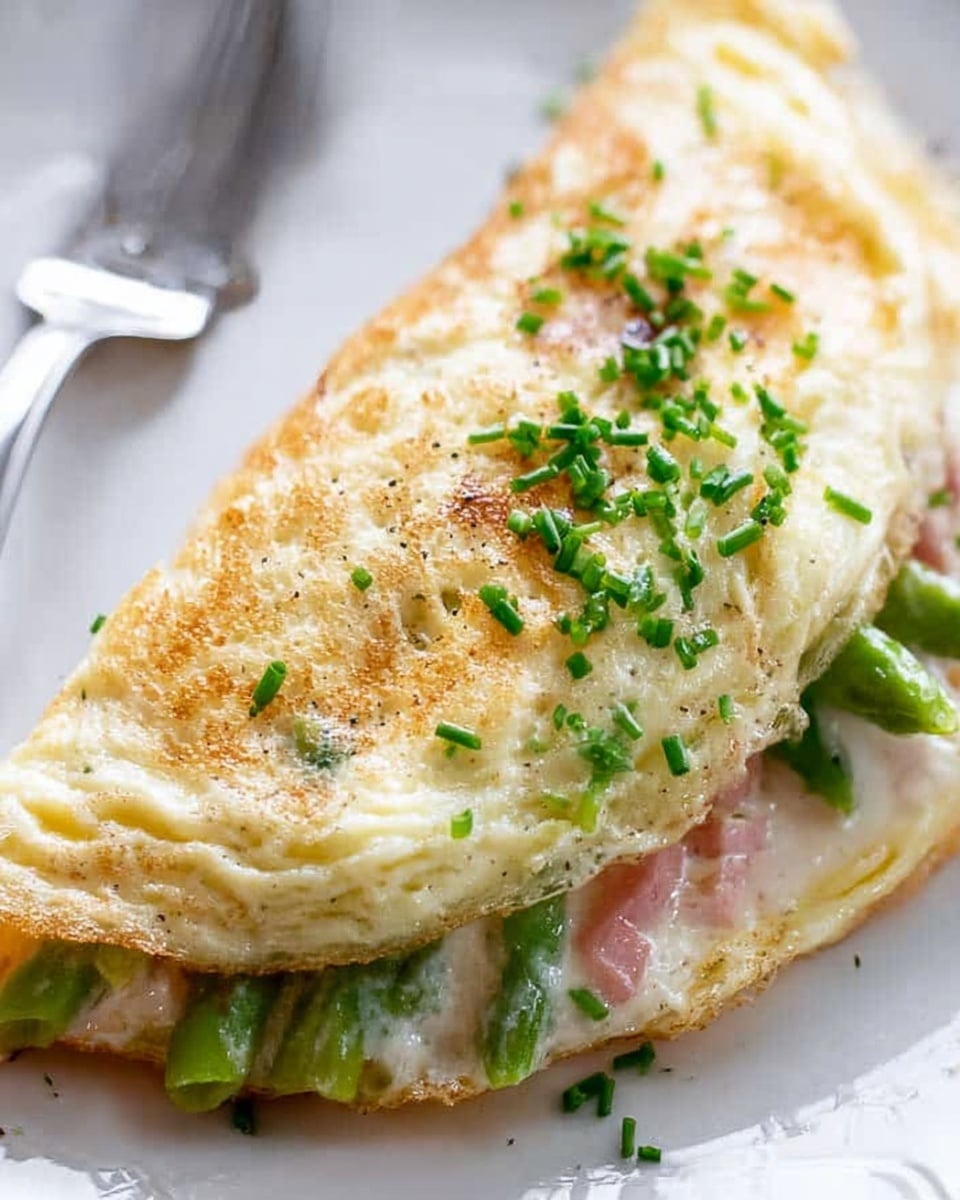 A fluffy white omelette folded in half sits on a white plate with a white marbled surface in the background. The top layer of the omelette is light golden with small scattered green chives. Inside, you see bright green asparagus and pink slices of ham peeking out from the soft, white egg texture. The edges of the omelette are slightly browned. A silver fork rests on the plate's edge near the omelette. photo taken with an iphone --ar 4:5 --v 7