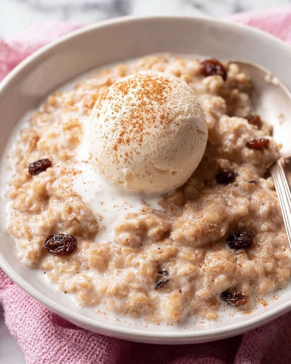 A close-up image of a white bowl filled with creamy oatmeal that has visible pieces of raisins mixed throughout. The oatmeal is light brown and looks thick and soft, with a smooth texture. On top, there is a scoop of white ice cream or cream, sprinkled lightly with light brown cinnamon powder. A silver spoon is partly buried in the oatmeal on the right side of the bowl. The bowl is set on a light pink cloth, all placed on a white marbled surface. photo taken with an iphone --ar 4:5 --v 7