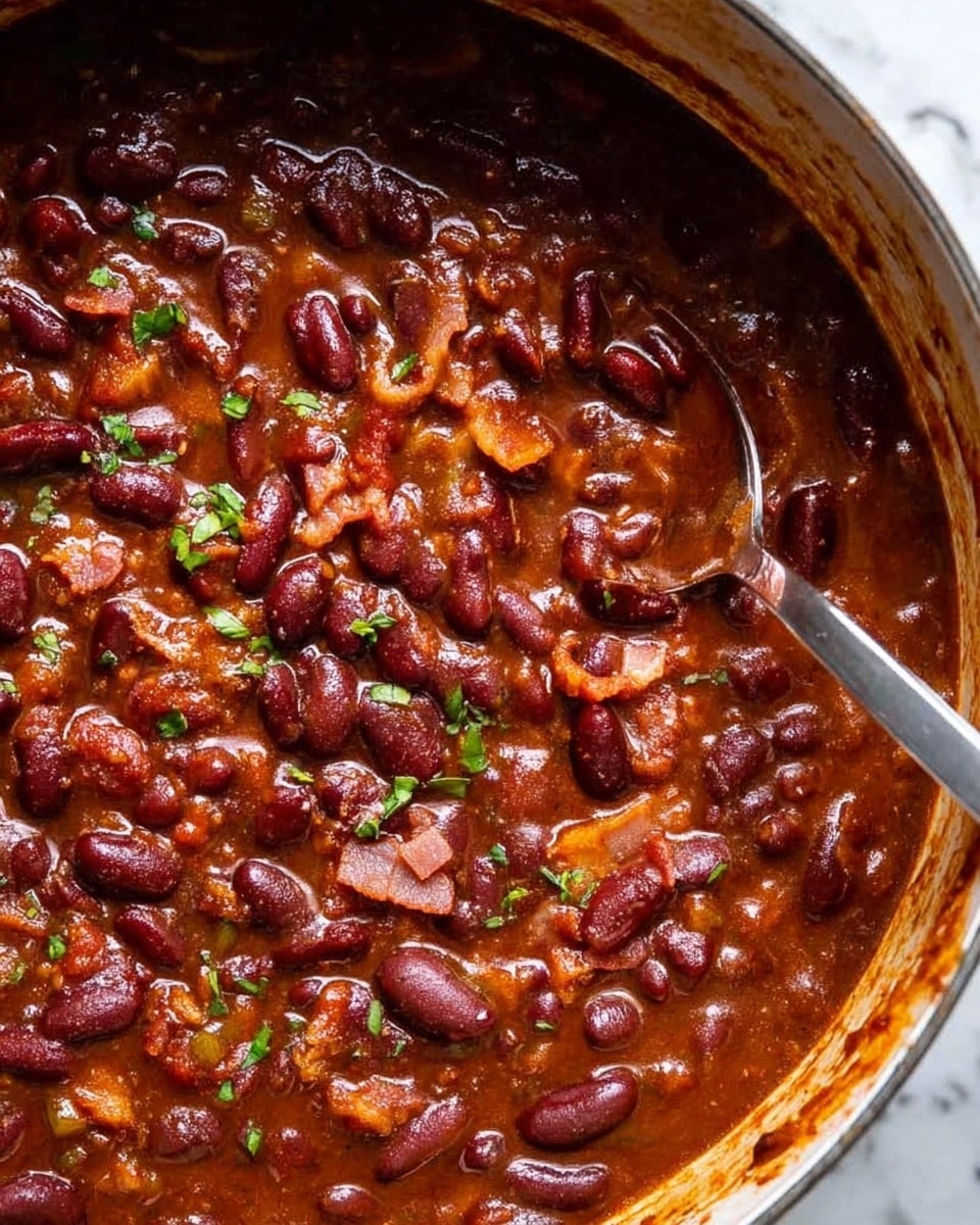 The image shows a close-up of a thick chili with dark red kidney beans scattered throughout a rich, deep brown sauce. Small pieces of crispy, browned bacon and tiny green herb bits are spread on top, adding texture and color contrast. The chili appears chunky and hearty with a glossy finish, all inside a pot with some sauce splattered on the sides. A metal spoon rests inside the pot, partially submerged in the chili. The photo is taken with an iphone --ar 4:5 --v 7