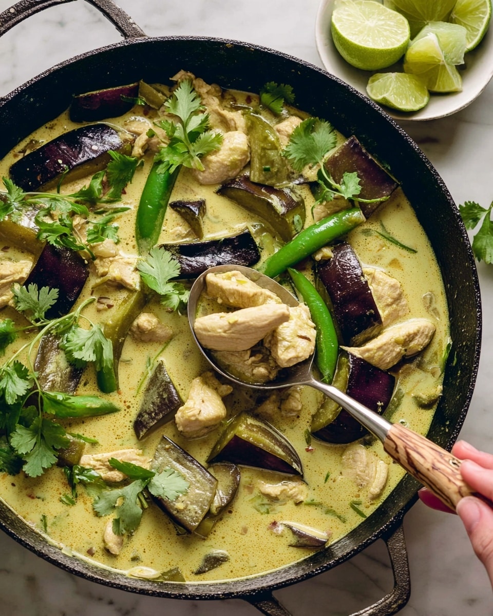 A close-up of a black cast iron pan filled with creamy green curry containing light brown chicken pieces, thick slices of dark purple eggplant, and whole green chili peppers. The curry sauce is light greenish-yellow and covers the ingredients smoothly. Bright green fresh cilantro leaves sit on top as garnish, adding a fresh pop of color. A large spoon with a wooden handle is partially submerged in the curry, lifting some chicken and a chili pepper. Next to the pan, a white bowl holds fresh lime wedges with a few cilantro leaves visible underneath. The whole scene rests on a white marbled surface. photo taken with an iphone --ar 4:5 --v 7