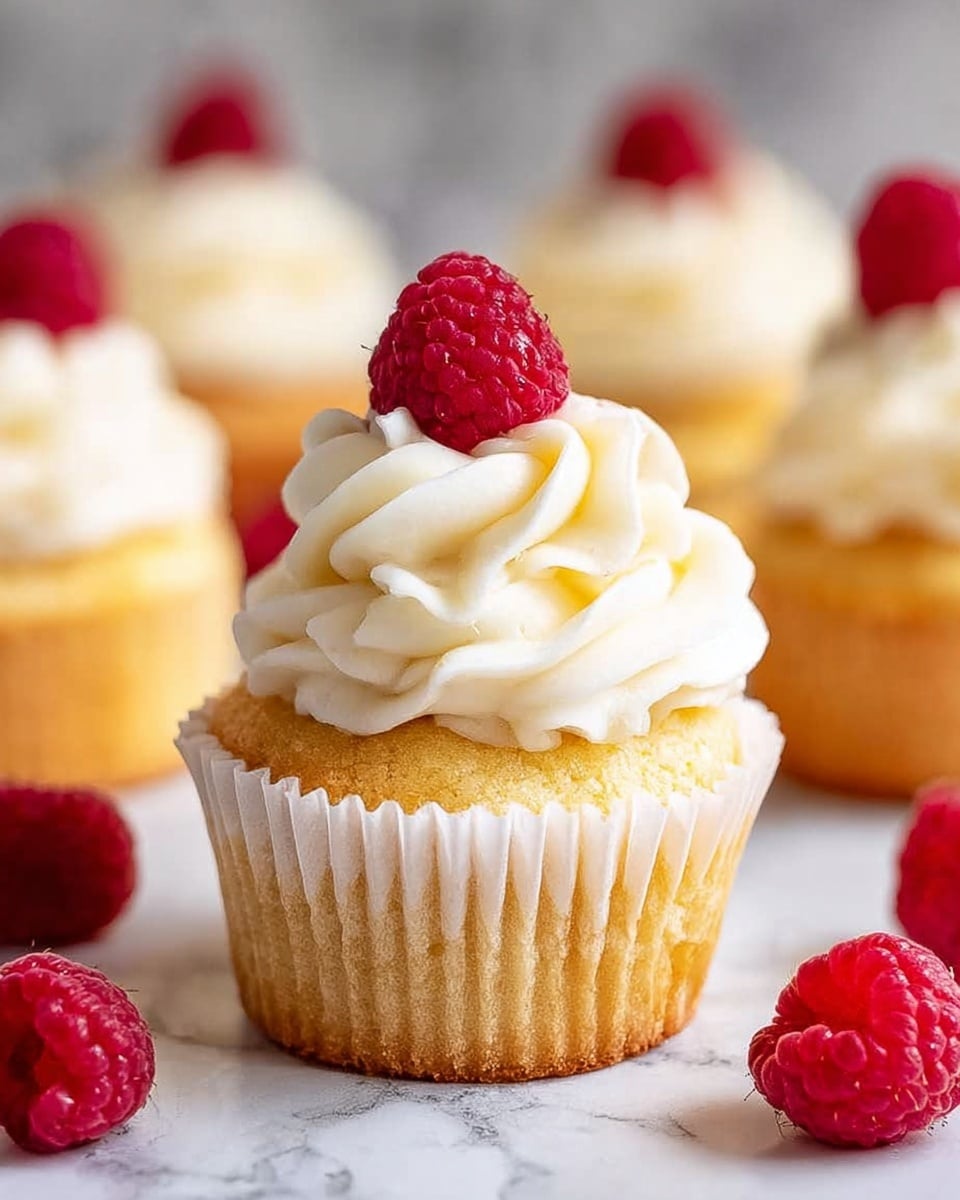 A single golden yellow cupcake sits on a white marbled surface with its white paper wrapper partially peeled back, showing a soft, crumbly texture. On top of the cupcake is a large swirl of smooth, creamy, white frosting, piled high in three thick, curvy layers. A bright red raspberry with a bumpy surface crowns the frosting. Around the cupcake are several more red raspberries scattered on the white marbled surface, and in the blurred background, another cupcake with similar frosting is visible. photo taken with an iphone --ar 4:5 --v 7