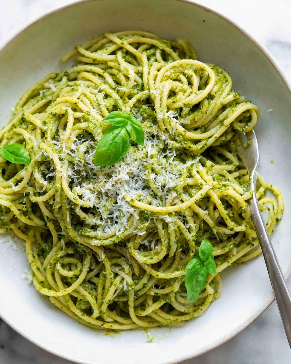 A close-up view of a plate filled with spaghetti covered in a green pesto sauce, the noodles are intertwined and lightly glossy. There are small green basil leaves scattered on top and finely grated white cheese sprinkled in the center. The plate is white and rests on a white marbled surface. A silver fork is partially visible on the right side of the plate. Photo taken with an iphone --ar 4:5 --v 7