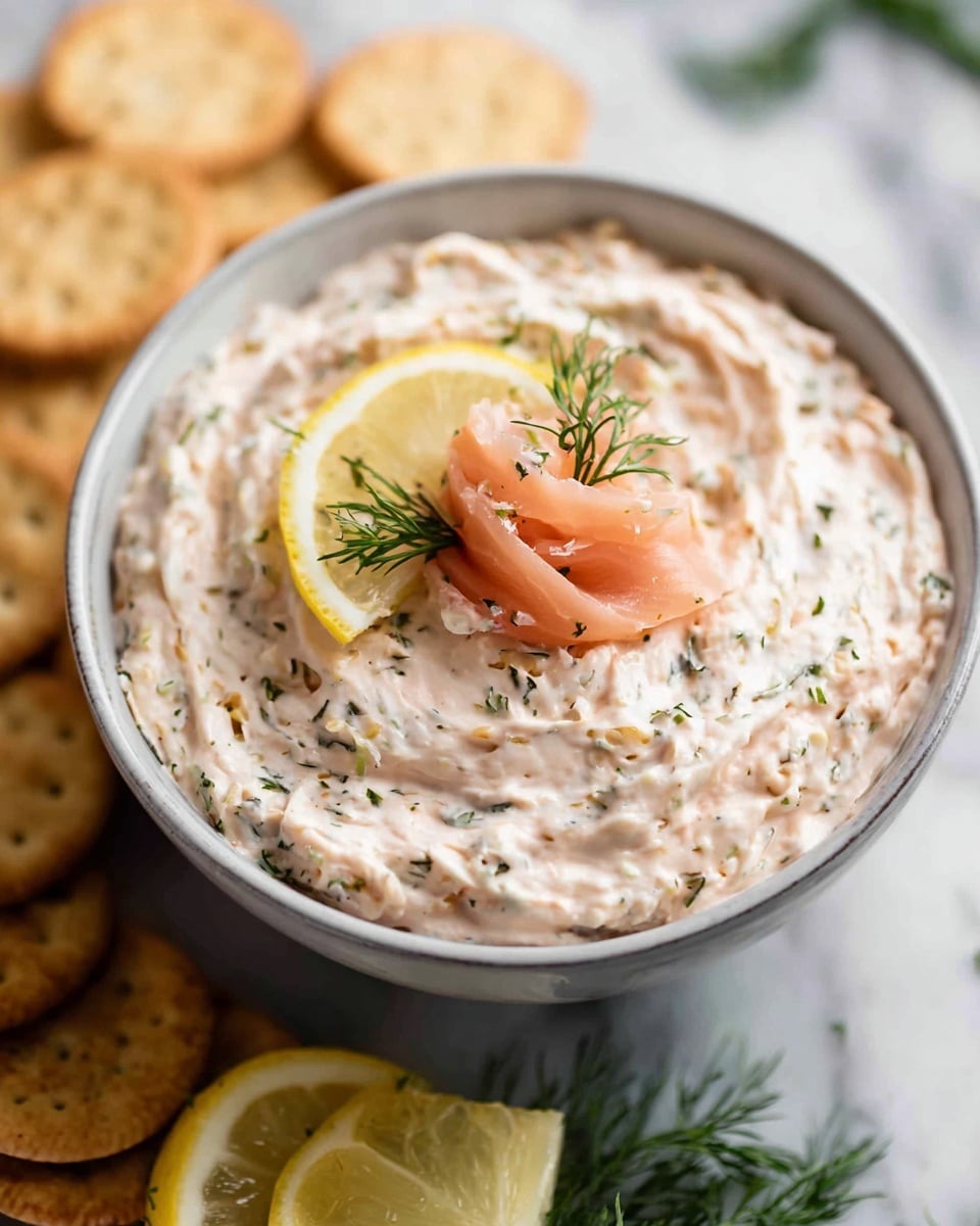 A close-up view of a bowl filled with a creamy, light beige dip mixed with small green herb pieces, swirled smoothly in three soft layers within the bowl. The dip is topped with a delicate rose-shaped piece of pink smoked salmon, two lemon slices with bright yellow skin and pale yellow fruit placed on the right edge, and garnished with fresh green dill sprigs scattered on top. The bowl is set on a white marbled surface with a few round beige crackers stacked slightly out of focus in the background. photo taken with an iphone --ar 4:5 --v 7