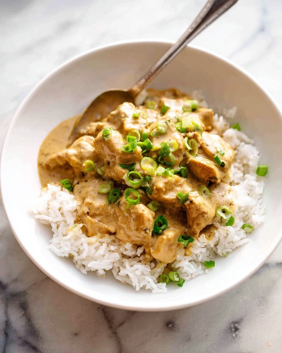 A white bowl filled with a base layer of fluffy white rice, topped with a thick, creamy beige sauce that covers tender pieces of chicken. The sauce has a smooth texture with a slight shine, and scattered on top are small slices of fresh green onions adding a pop of bright green color. A silver spoon rests inside the bowl on the right side, partially dipped into the sauce. The bowl sits on a white marbled surface with soft, natural lighting highlighting the dish’s warm and inviting look. Photo taken with an iphone --ar 4:5 --v 7