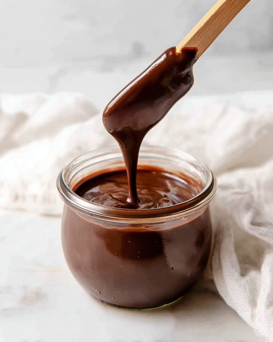 A clear glass jar filled with thick, smooth, dark brown chocolate sauce sits on a white marbled surface. A wooden spreader holds a generous amount of the glossy chocolate sauce dripping slowly back into the jar, showing the sauce's creamy texture and rich color. Soft, diffused light highlights the shiny surface of the sauce and the reflection on the jar edges, creating a warm and inviting scene. Photo taken with an iphone --ar 4:5 --v 7